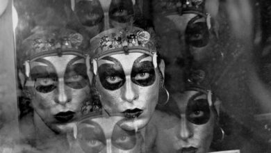 A imagem apresenta um retrato em preto e branco de uma pessoa com maquiagem dramática, incluindo olhos destacados em preto e lábios escuros. A figura parece refletida em uma superfície, criando um efeito de múltiplas camadas. O fundo é desfocado, e há um leve brilho que sugere um ambiente de backstage ou de performance.