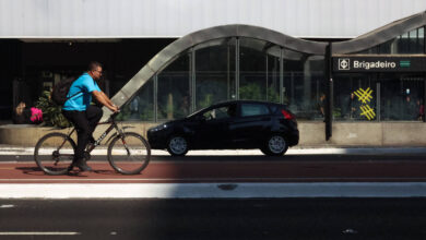 A imagem mostra um ciclista pedalando em uma ciclovia, ao lado de uma rua onde um carro preto está estacionado. Ao fundo, há uma estrutura arquitetônica com um design curvo e moderno, e uma estação de metrô com o nome