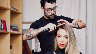 A imagem mostra um cabeleireiro estilizando o cabelo de uma cliente. O cabeleireiro é um homem com óculos e barba, usando uma camisa preta. A cliente é uma mulher com cabelo longo e liso, vestindo um vestido preto. O ambiente parece ser um salão de beleza, com prateleiras ao fundo e uma cadeira de cabeleireiro. Há uma luz suave iluminando a cena.