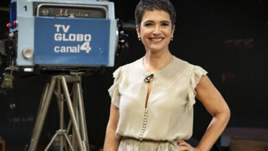 Uma mulher sorridente está em pé em um estúdio de televisão, vestindo um vestido claro. Ao fundo, há uma câmera de TV antiga com a inscrição