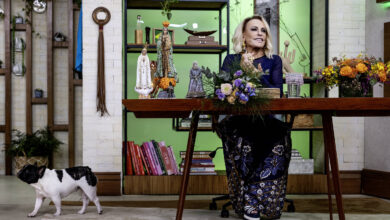 Uma apresentadora está sentada em uma mesa de madeira em um estúdio decorado. Ela usa um vestido azul escuro com detalhes em dourado e está cercada por arranjos de flores. Ao fundo, há prateleiras com livros e objetos decorativos. Um cão de pequeno porte, com pelagem branca e manchas pretas, caminha pelo chão ao lado da mesa.