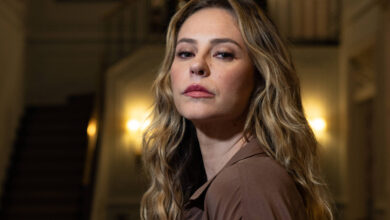 A imagem mostra uma mulher com cabelo longo e ondulado, usando uma blusa marrom. Ela está em um ambiente interno elegante, com uma escada ao fundo e um lustre iluminado. A mulher está em um ângulo ligeiramente de perfil, com uma expressão séria e as mãos unidas à frente do corpo.