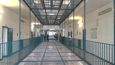 A imagem mostra um corredor de prisão com paredes brancas e um piso de metal. O corredor é cercado por grades metálicas e possui portas em ambos os lados. Há iluminação artificial nas paredes e uma grade no teto, permitindo a entrada de luz natural. O ambiente parece limpo e organizado, com um espaço amplo e sem pessoas visíveis.