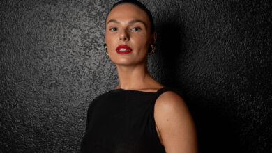 A imagem mostra uma mulher com cabelo preso e maquiagem marcante, incluindo batom vermelho. Ela está vestindo uma blusa preta e posando em frente a um fundo texturizado e escuro. A iluminação destaca seu rosto e expressões faciais.