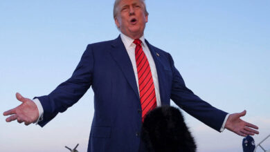 Imagem mostra Donald Trump, um homem em traje formal, com um terno azul e uma gravata vermelha, está de pé em uma pista de pouso, com os braços abertos e falando. Ele parece estar em uma entrevista ou discurso. Ao fundo, há uma pista de avião e algumas pessoas em trajes formais. Microfones estão posicionados próximos a ele, e o céu está claro.