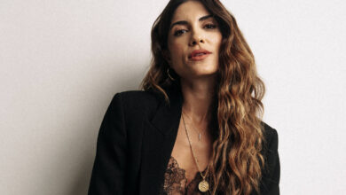 A imagem mostra uma mulher posando em pé, vestindo um blazer preto sobre uma blusa marrom com detalhes em renda. Ela tem cabelos longos e ondulados, e está usando acessórios como colares. O fundo é claro e neutro, destacando a figura da mulher.