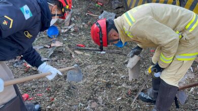 A imagem mostra dois homens trabalhando em uma área de destroços. Um deles está vestido com um uniforme de bombeiro, enquanto o outro usa um uniforme de polícia. Ambos estão usando capacetes vermelhos e estão agachados, utilizando ferramentas de escavação, como pás, para remover detritos do chão. O ambiente parece ser um local de acidente ou deslizamento, com vários pedaços de materiais espalhados ao redor.