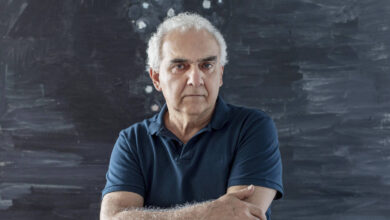 Um homem de cabelos grisalhos e pele clara está em pé, com os braços cruzados, em frente a uma parede de cor escura com manchas brancas. Ele usa uma camiseta azul escura e tem uma expressão séria. O fundo parece ser uma obra de arte abstrata, com uma textura irregular e cores escuras.